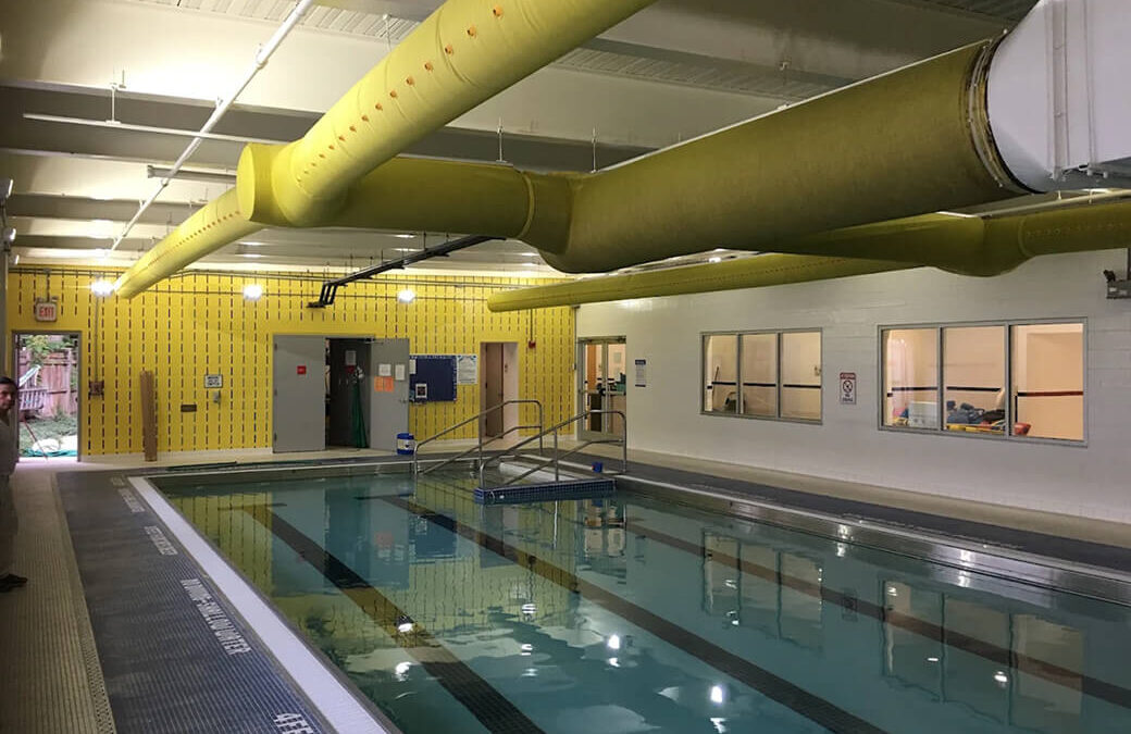 Indoor commercial pool area with painted white walls, gray ceiling beams and bright yellow ventilation ducts.