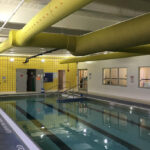 Indoor commercial pool area with painted white walls, gray ceiling beams and bright yellow ventilation ducts.