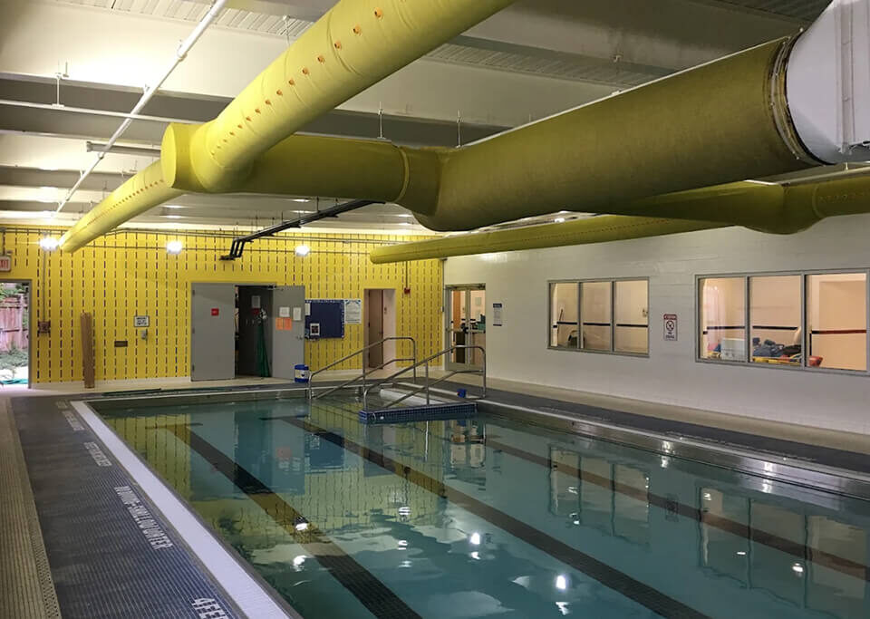 Indoor commercial pool area with painted white walls, gray ceiling beams and bright yellow ventilation ducts.
